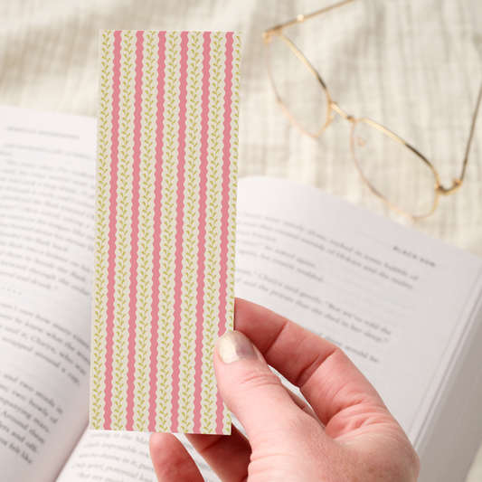 Hand holding a striped pink and yellow bookmark over an open book with glasses in the background.