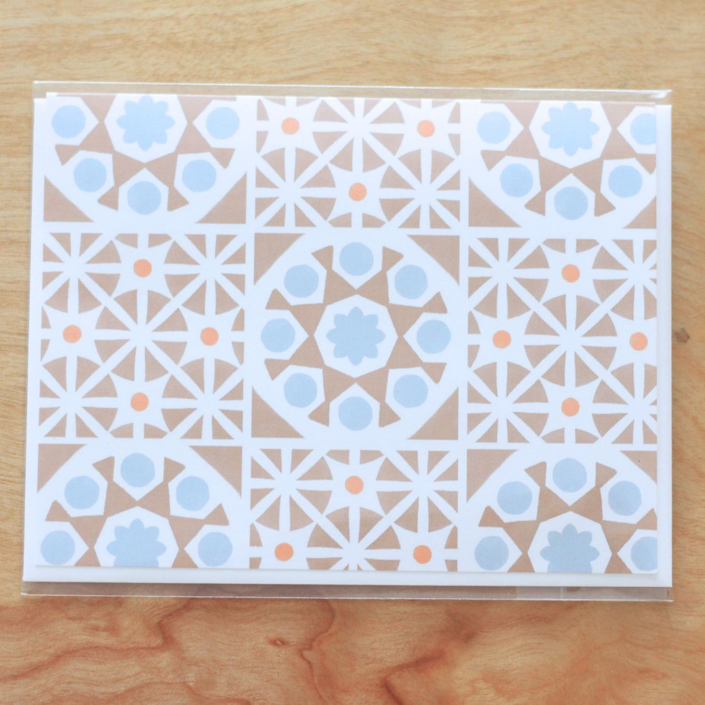 A horizontal greeting card featuring an all-over pattern of tan-colored Mediterranean tiles arranged in a symmetrical grid format. The folded card is shown stacked on a white envelope. The card is inside clear cello packaging and is centered against a wood background.