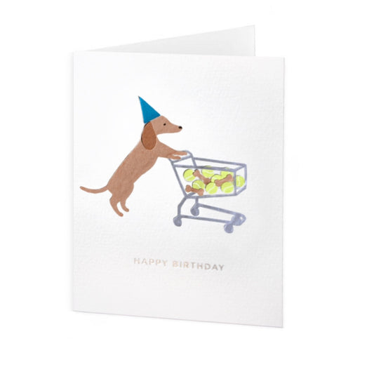 Folded greeting card standing up vertically with a dachshund in a blue party hat pushing a shopping cart full of brown dog treats and tennis balls. Under the dog is silver-colored phrase "Happy Birthday" in foil embossed all caps font. The greeting card design is centered on a white textured card background.
