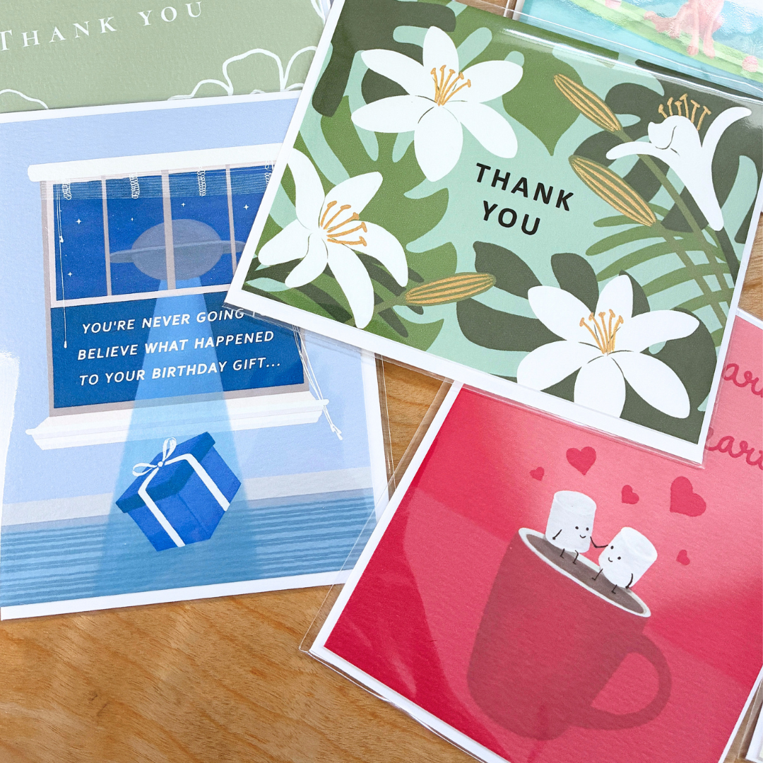 an assortment of illustrated greeting cards, individually wrapped in clear protective sleeves, are casually layered on a light-colored wood grain table. the central card features a lush, botanical "thank you" design with large white lilies and tropical monstera and palm leaves against a sage green background. To its left, a whimsical blue birthday card shows a ufo outside a window beaming up a wrapped present with the text, "you're never going to believe what happened to your birthday gift..." 