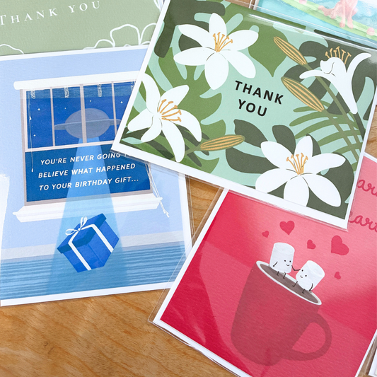 an assortment of illustrated greeting cards, individually wrapped in clear protective sleeves, are casually layered on a light-colored wood grain table. the central card features a lush, botanical "thank you" design with large white lilies and tropical monstera and palm leaves against a sage green background. To its left, a whimsical blue birthday card shows a ufo outside a window beaming up a wrapped present with the text, "you're never going to believe what happened to your birthday gift..." 