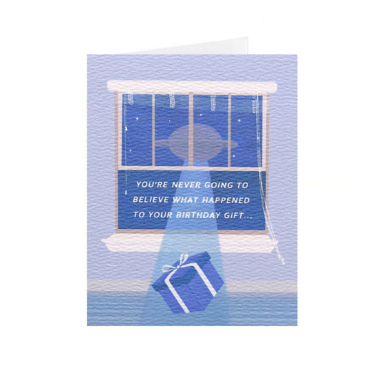 A vertical birthday card stands up on a centered white background. The card has an illustration of a blue wrapped present being lifted out of the open light blue window to a floating UFO. The white text  centered in the open window says, 'you're never going to believe what happened to your birthday gift...' In the night sky there are small stars.