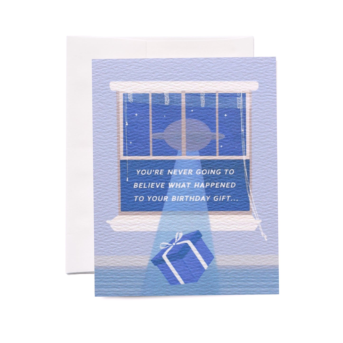 A vertical folded greeting card  laying flat with an envelope tucked behind it, centered on a white background. The card has an illustration of a blue wrapped present being lifted out of the open light blue window to a floating UFO. The white text centered in the open window says, 'you're never going to believe what happened to your birthday gift...' In the night sky there are small stars.