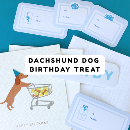 Birthday card with a dachshund pushing a shopping cart, a “Howdy” sticker, and stacked “To & From” gift sticker labels, with a white envelope stacked under the card and a folded typed letter under the envelope.