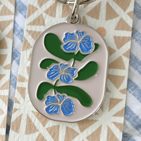 An oval-shaped silver enamel keychain features a stylized botanical design with three blue flowers with fine silver-colored detailing are stacked vertically along a central stem, interspersed with broad, rounded green leaves. The keychain is displayed on a backing card with a tan geometric triangle pattern, all resting on a soft blue and white striped fabric.
