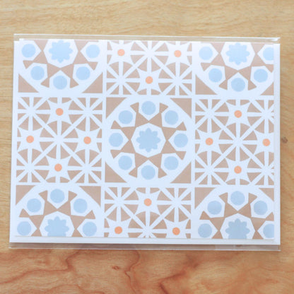 A horizontal greeting card featuring an all-over pattern of tan-colored Mediterranean tiles arranged in a symmetrical grid format. The folded card is shown stacked on a white envelope. The card is inside clear cello packaging and is centered against a wood background.