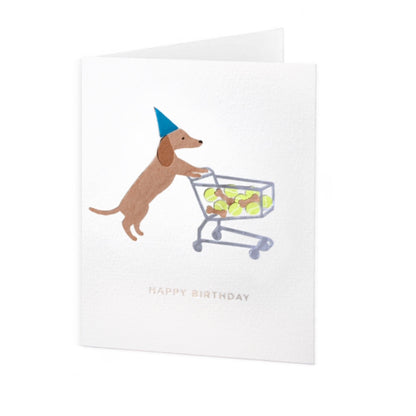 Folded greeting card standing up vertically with a dachshund in a blue party hat pushing a shopping cart full of brown dog treats and tennis balls. Under the dog is silver-colored phrase "Happy Birthday" in foil embossed all caps font. The greeting card design is centered on a white textured card background.