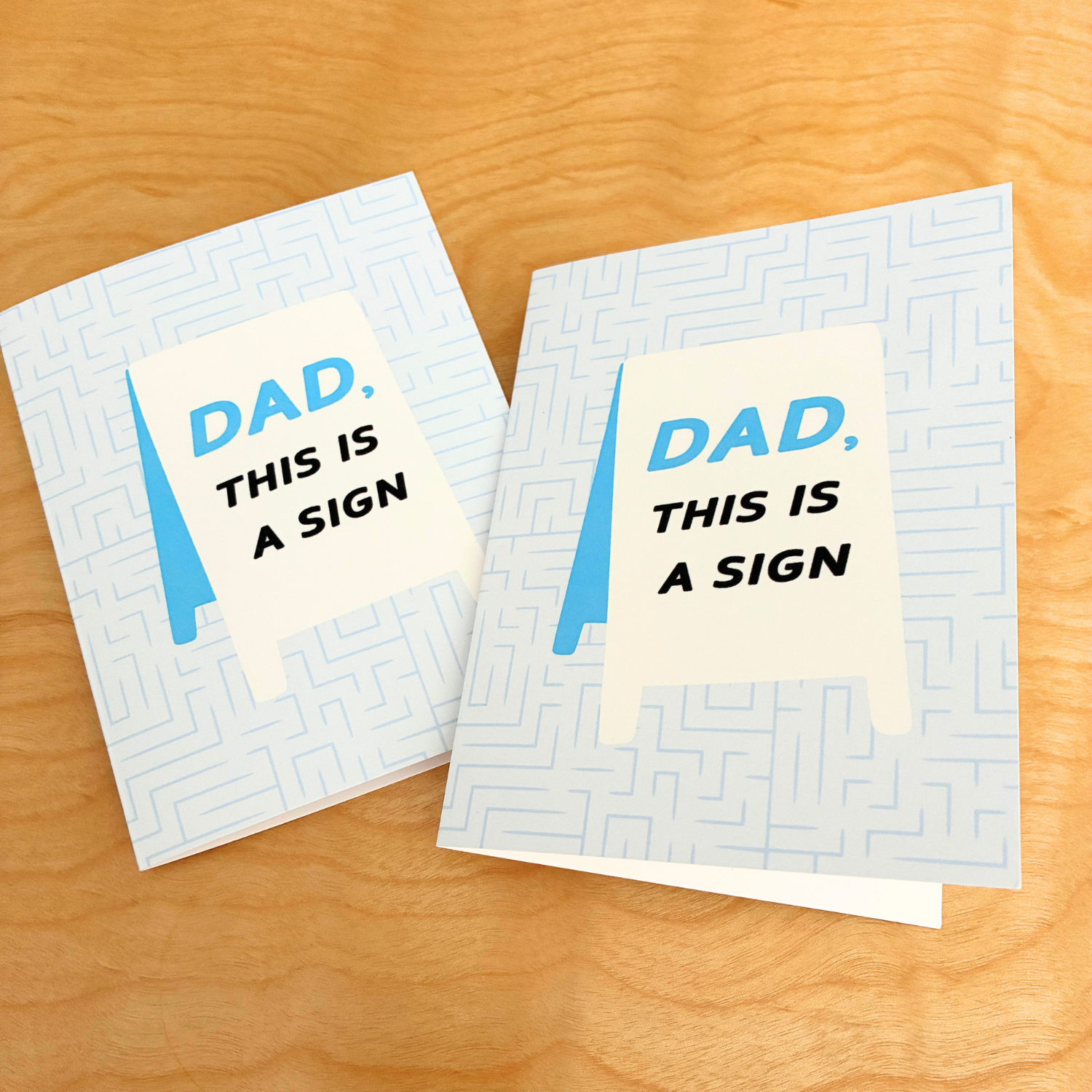 Two identical folded greeting cards sit side-by-side on a light-colored wooden table with a visible grain. The cards have a light blue background filled with a subtle, geometric maze pattern. On the front of each card is an illustration of a white A-frame sidewalk sign. The text printed on the sign reads: "Dad," in blue, followed by "This is a sign" in bold black capital letters. The card on the left is angled slightly upward, while the card on the right is upright and slightly open.