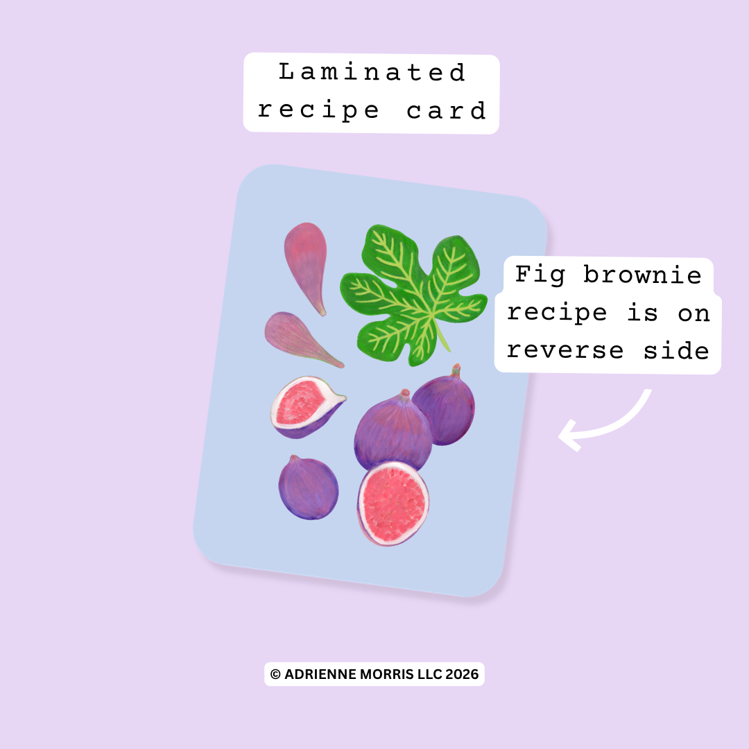 A laminated recipe card featuring illustrated figs and leaves on a light blue background, shown on a lavender backdrop with text labels reading 'Laminated recipe card' and 'Fig brownie recipe is on reverse side.'.