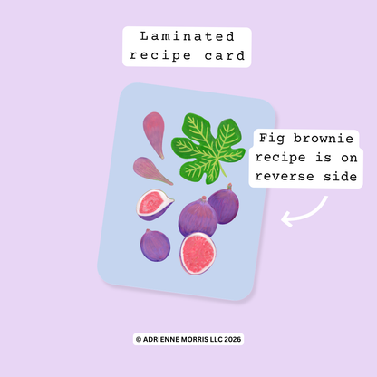 A laminated recipe card featuring illustrated figs and leaves on a light blue background, shown on a lavender backdrop with text labels reading 'Laminated recipe card' and 'Fig brownie recipe is on reverse side.'.