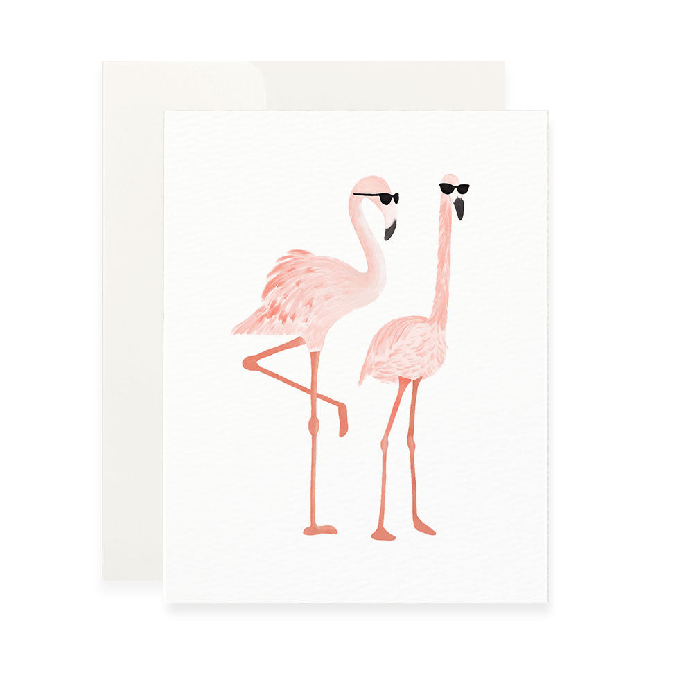A vertical white greeting card featuring a watercolor-style illustration of two pink flamingos wearing black sunglasses. The flamingo on the left stands on one leg, while the one on the right stands on both. A plain white envelope is tucked behind the card.