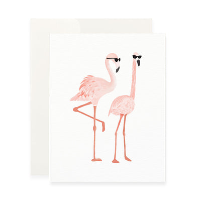 A vertical white greeting card featuring a watercolor-style illustration of two pink flamingos wearing black sunglasses. The flamingo on the left stands on one leg, while the one on the right stands on both. A plain white envelope is tucked behind the card.