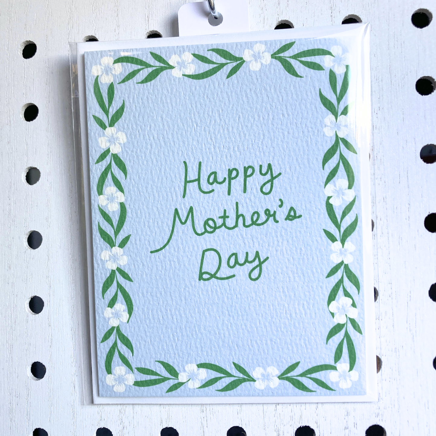 A light-blue textured greeting card with a hand-lettered message 'Happy Mother's Day' in green. The card has a hand-painted floral and vine border and is in a clear plastic sleeve, hanging from a metal pegboard hook on a white pegboard background.