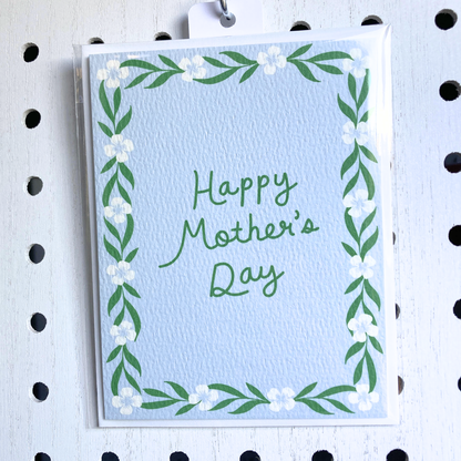 A light-blue textured greeting card with a hand-lettered message 'Happy Mother's Day' in green. The card has a hand-painted floral and vine border and is in a clear plastic sleeve, hanging from a metal pegboard hook on a white pegboard background.