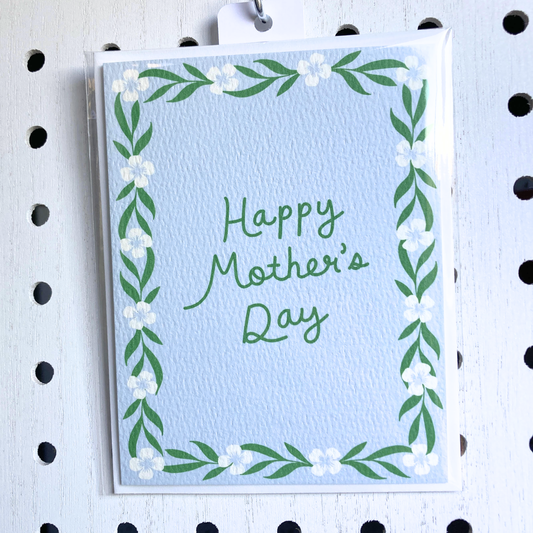 A light-blue textured greeting card with a hand-lettered message 'Happy Mother's Day' in green. The card has a hand-painted floral and vine border and is in a clear plastic sleeve, hanging from a metal pegboard hook on a white pegboard background.