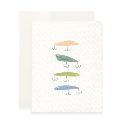 This vertical greeting card features a clean, minimalist design centered on a piece of white, textured paper. The artwork consists of illustrated fishing lures stacked vertically in the center. Starting from the top, the first lure is a muted yellow color facing right, followed by a sage green lure facing left, then a bright olive green lure facing right, and finally a slate blue lure facing left at the bottom. Two shiny silver-grey treble hooks hang under each lure. 