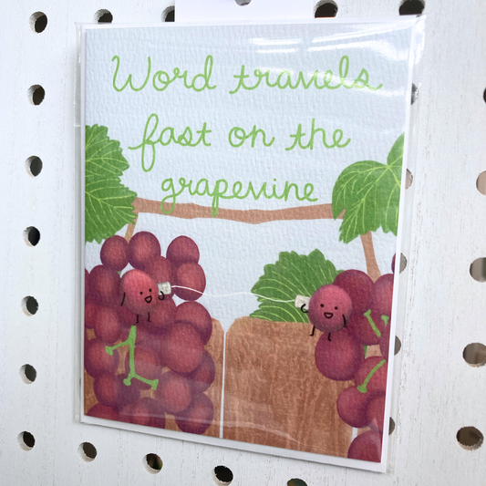 A vertical folded greeting card features a playful illustration of two red grape clusters separated by a wooden fence against a soft blue sky. In the center, two personified grapes with tiny smiling faces and thin black limbs "talk" to each other using a classic tin-can telephone connected by a white string. The top of the card displays the phrase "Word travels fast on the grapevine" in a friendly, green cursive font. The artwork is on textured paper and shown packaged in a clear bag
