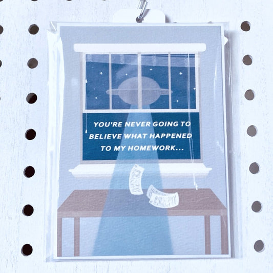 A wrapped greeting card on a white pegboard. The card is light blue with a white envelope tucked behind it. A light blue text box in the middle of the card shows white text. The card has an illustration of a brown table with three white sheets of paper levitating out of the open light blue window to a floating UFO. The text on the light blue box says, 'you're never going to believe what happened to my homework...' At the end of the text is five small stars.