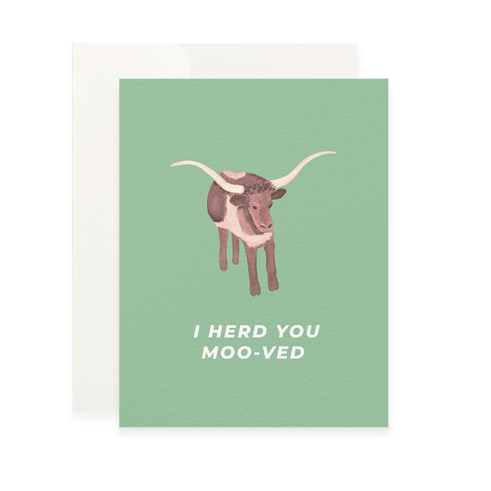 a light green greeting card featuring a painted illustration of a brown and white longhorn cow. below the cow, the white sans-serif text reads, "i herd you moo-ved." the card is shown with a plain white envelope behind it.