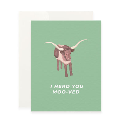 a light green greeting card featuring a painted illustration of a brown and white longhorn cow. below the cow, the white sans-serif text reads, "i herd you moo-ved." the card is shown with a plain white envelope behind it.