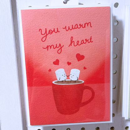 Large greeting card packaged in clear bag hanging on metal s-hook on  a white peg board. The card is packaged with one off-white envelope. The card is red with cursive red lettering that says "you warm my heart" with art illustration of two marshmallow people holding hands sitting on a steaming mug.