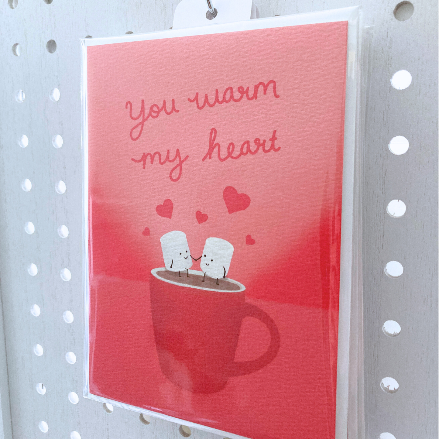 Large greeting card packaged in clear bag hanging on metal s-hook on  a white peg board. The card is packaged with one off-white envelope. The card is red with cursive red lettering that says "you warm my heart" with art illustration of two marshmallow people holding hands sitting on a steaming mug.