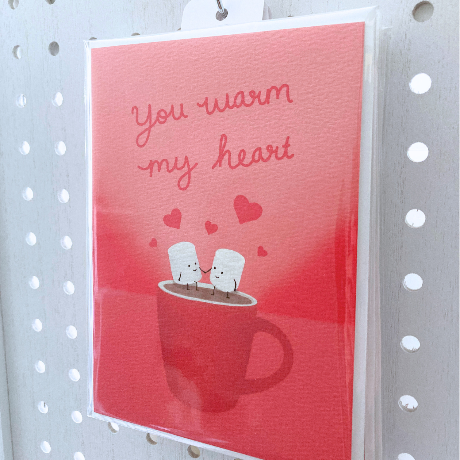 Large greeting card packaged in clear bag hanging on metal s-hook on  a white peg board. The card is packaged with one off-white envelope. The card is red with cursive red lettering that says "you warm my heart" with art illustration of two marshmallow people holding hands sitting on a steaming mug.