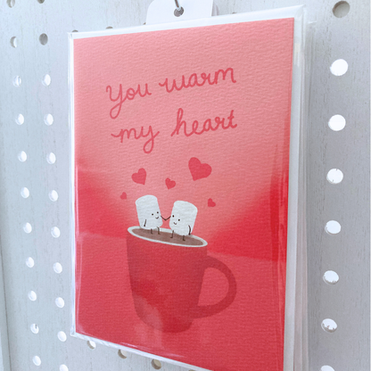 Large greeting card packaged in clear bag hanging on metal s-hook on  a white peg board. The card is packaged with one off-white envelope. The card is red with cursive red lettering that says "you warm my heart" with art illustration of two marshmallow people holding hands sitting on a steaming mug.