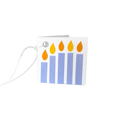 Paper gift tag with five paper candles in purple each topped with a yellow flame. The gift tag is attached by a white string.