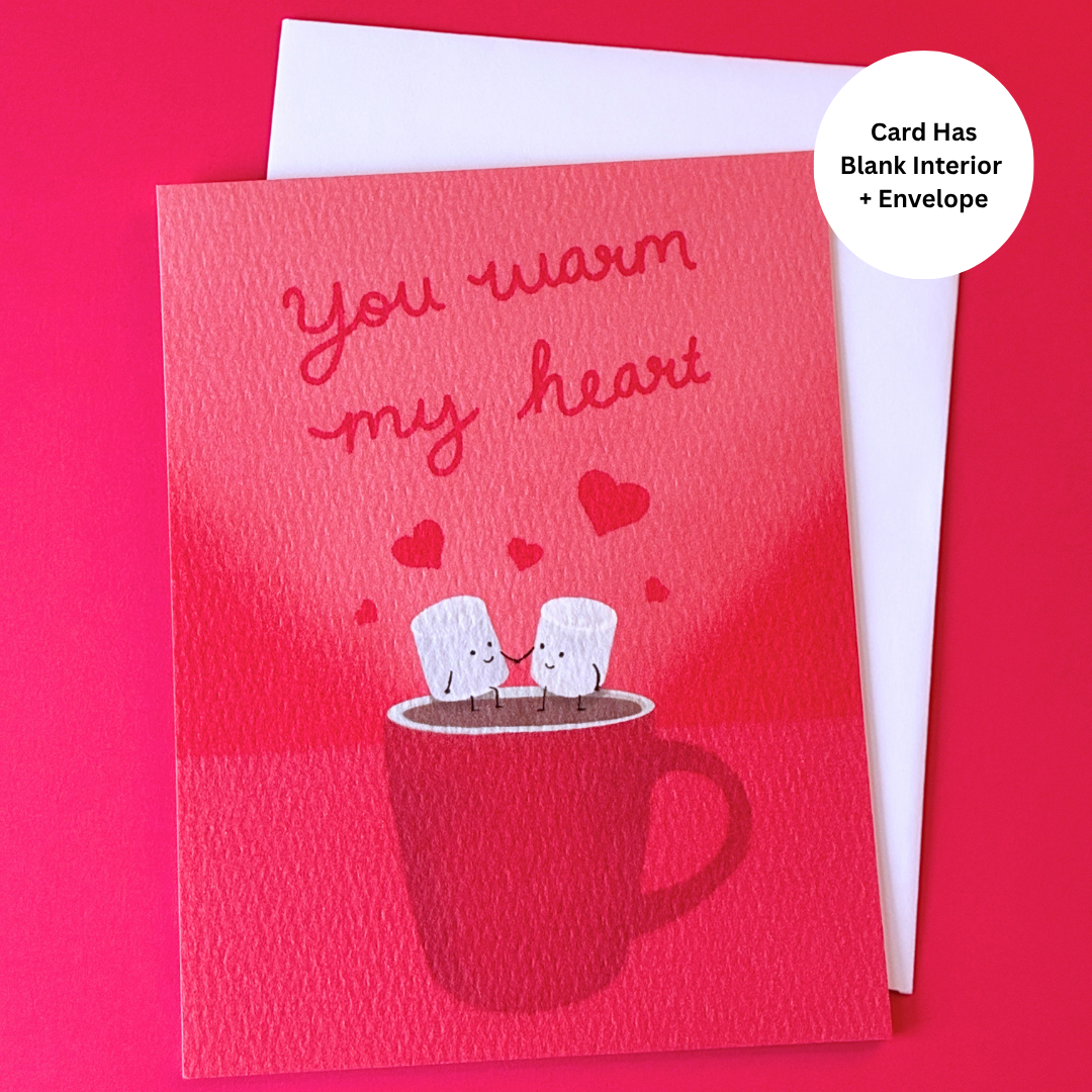 A red greeting card with the phrase 'you warm my heart' above an illustration of two marshmallows sitting on a mug, shown with a white envelope behind it on a bright pink background. A white circular badge says 'Card Has Blank Interior + Envelope'.