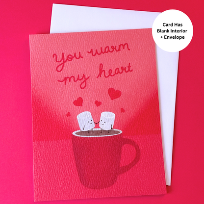 A red greeting card with the phrase 'you warm my heart' above an illustration of two marshmallows sitting on a mug, shown with a white envelope behind it on a bright pink background. A white circular badge says 'Card Has Blank Interior + Envelope'.