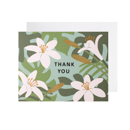 A folded horizontal thank you card resting flat on a white surface with a white envelope tucked underneath. The card features an illustrated pattern of white lilies and green tropical leaves against a light teal-blue background. The words "thank you" are centered in black capital letters. Some floral elements are accented with metallic gold-colored ink.