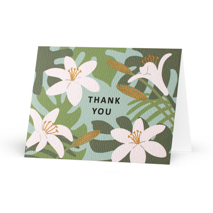 A folded horizontal thank you card standing up on a white surface. The card features an illustrated pattern of white lilies and green tropical leaves against a light teal-blue background. The words "thank you" are centered in black capital letters. Some floral elements are accented with metallic gold-colored ink.