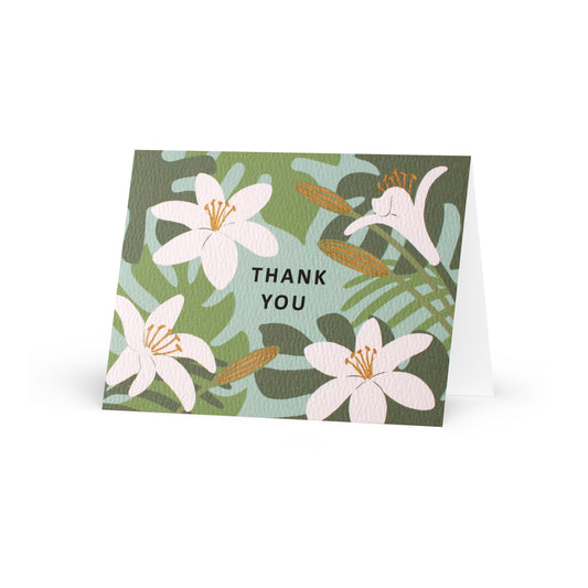 A folded horizontal thank you card standing up on a white surface. The card features an illustrated pattern of white lilies and green tropical leaves against a light teal-blue background. The words "thank you" are centered in black capital letters. Some floral elements are accented with metallic gold-colored ink.