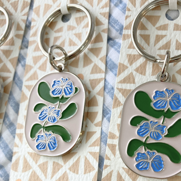 Two oval-shaped silver enamel keychains with stylized botanical designs. Each keychain has three blue flowers with fine silver-colored detailing are stacked vertically along a central stem, interspersed with broad, rounded green leaves. The keychain is displayed on a backing card with a tan geometric triangle pattern, all resting on a soft blue and white striped fabric.