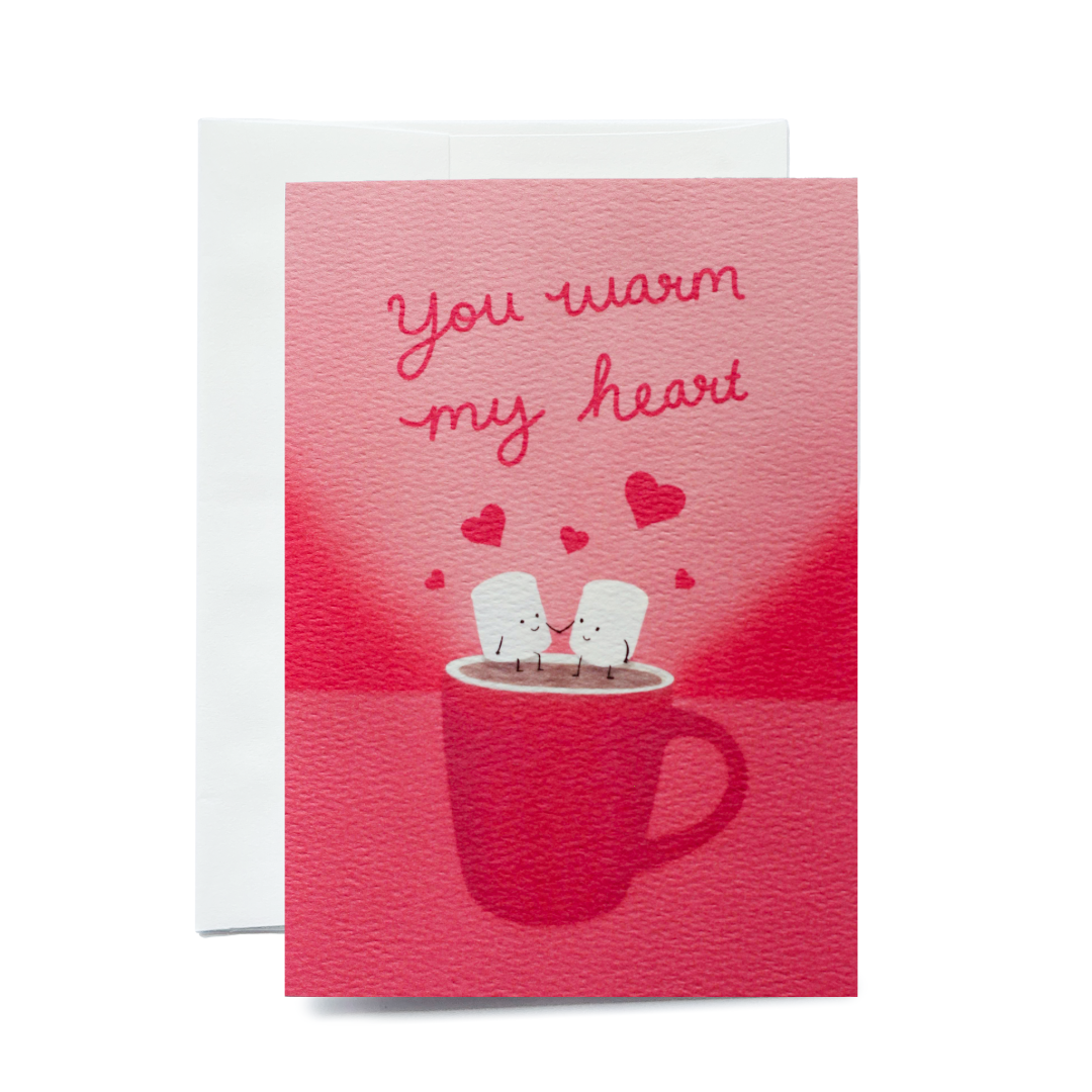 A vertical greeting card showing two personified, smiling marshmallows holding hands in a red mug of hot chocolate. The scene includes floating red hearts and a textured pink background. The card is shown with a white envelope on a plain white background.