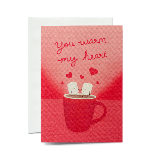 A vertical greeting card showing two personified, smiling marshmallows holding hands in a red mug of hot chocolate. The scene includes floating red hearts and a textured pink background. The card is shown with a white envelope on a plain white background.
