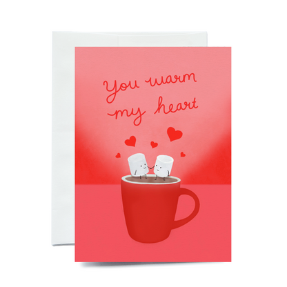 A vertical greeting card showing two personified, smiling marshmallows holding hands in a red mug of hot chocolate. The scene includes floating red hearts and a textured pink background. The card is shown with a white envelope on a plain white background.