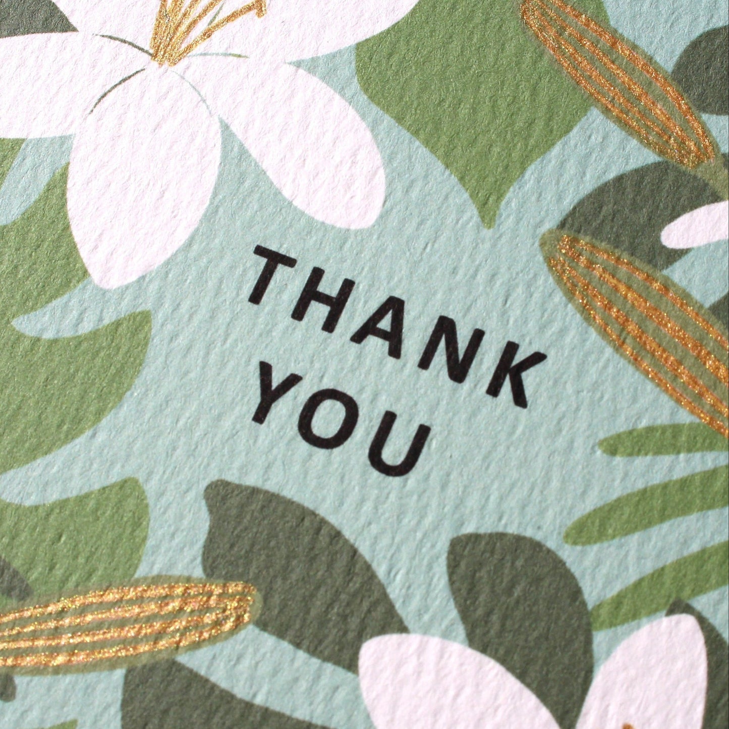 a close-up, angled view of a textured card featuring white lilies and green tropical leaves. the centers of the lilies and the elongated flower buds are detailed with hand-drawn shimmering gold ink. the words "thank you" are printed in black block letters in the center of the floral design
