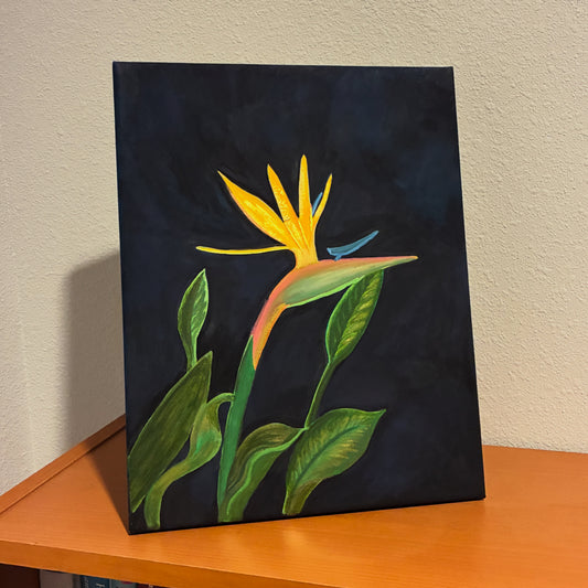 A vertical canvas painting of a yellow bird of paradise flower with green leaves on a dark navy background, leaning against a beige wall on an orange wooden table.