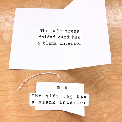 A folded white greeting card and a matching gift tag laid on a light wood surface. Both pieces of paper have black typewriter-style text reading, 'The palm trees folded card has a blank interior' and 'The gift tag has a blank interior' respectively.