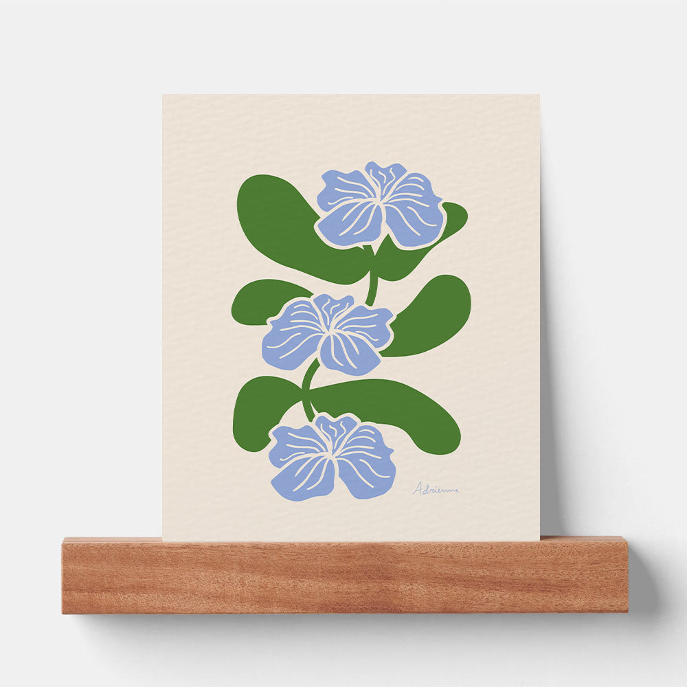 a vertical minimalist botanical art print resting on a walnut wood picture ledge against a plain white wall. the print features three stylized light blue flowers with white outlines, stacked vertically on a winding green stem with large rounded leaves against a cream textured background.
