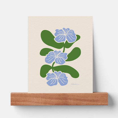 a vertical minimalist botanical art print resting on a walnut wood picture ledge against a plain white wall. the print features three stylized light blue flowers with white outlines, stacked vertically on a winding green stem with large rounded leaves against a cream textured background.