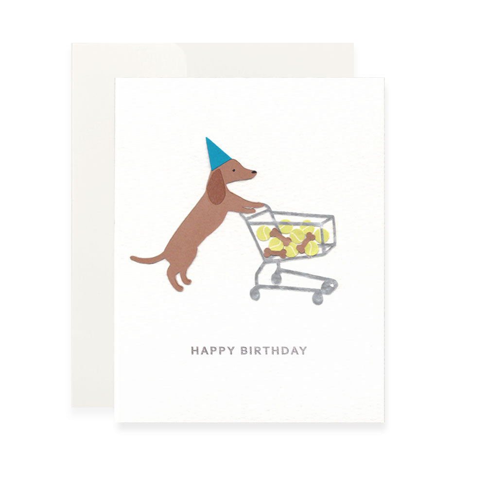 Cut paper art of a brown dachshund dog wearing a bright blue cone-shaped party hat. The dog is standing on its hind legs, pushing a silver wire shopping cart filled with several yellow tennis balls and brown dog bone treats. the words "happy birthday" are printed in silver sans-serif letters at the bottom of the card.