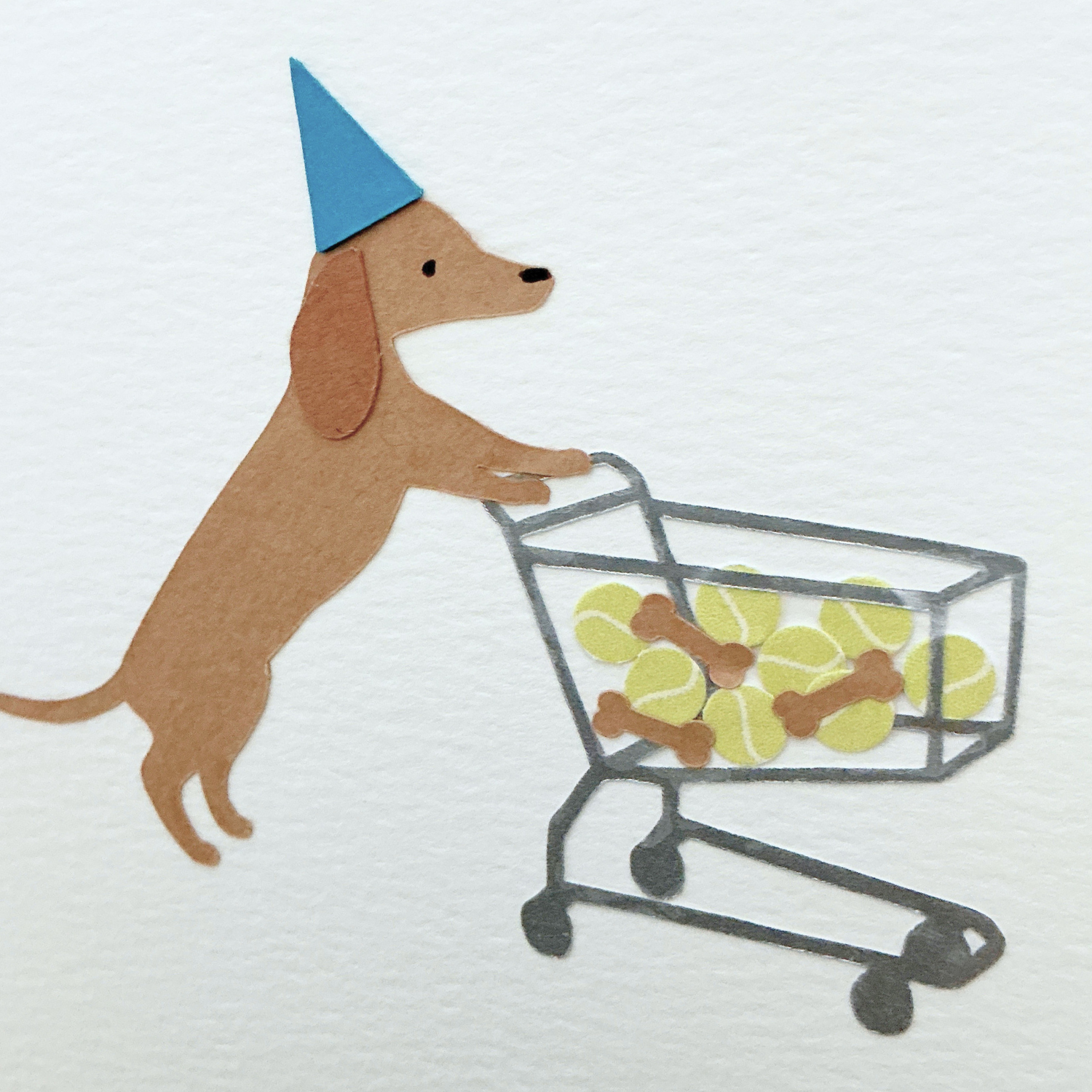 Paper Dog with a party hat pushing a shopping cart filled with tennis balls and dog bones on a white textured paper background.