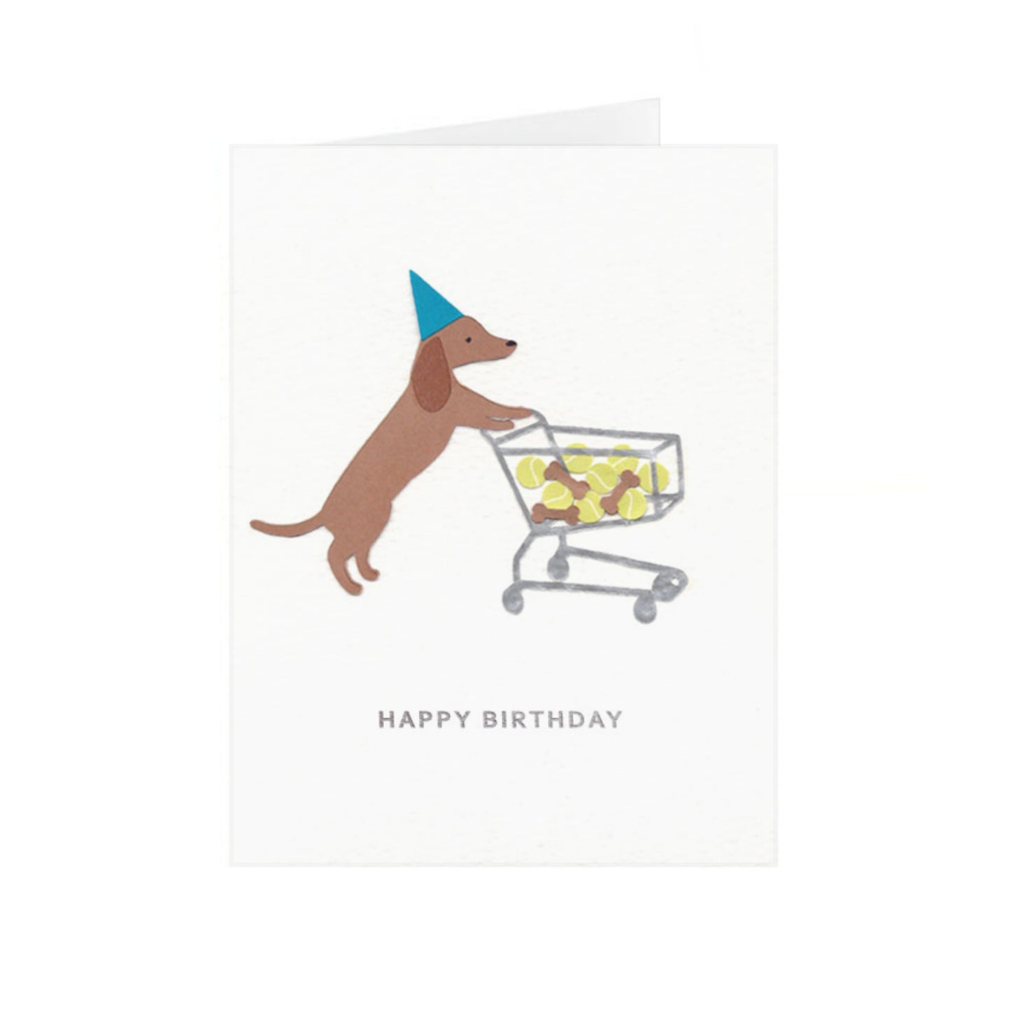 Folded greeting card standing up vertically with a dachshund in a blue party hat pushing a shopping cart full of brown dog treats and tennis balls. Under the dog is silver-colored phrase "Happy Birthday" in foil embossed all caps font. The greeting card design is centered on a white textured card background.