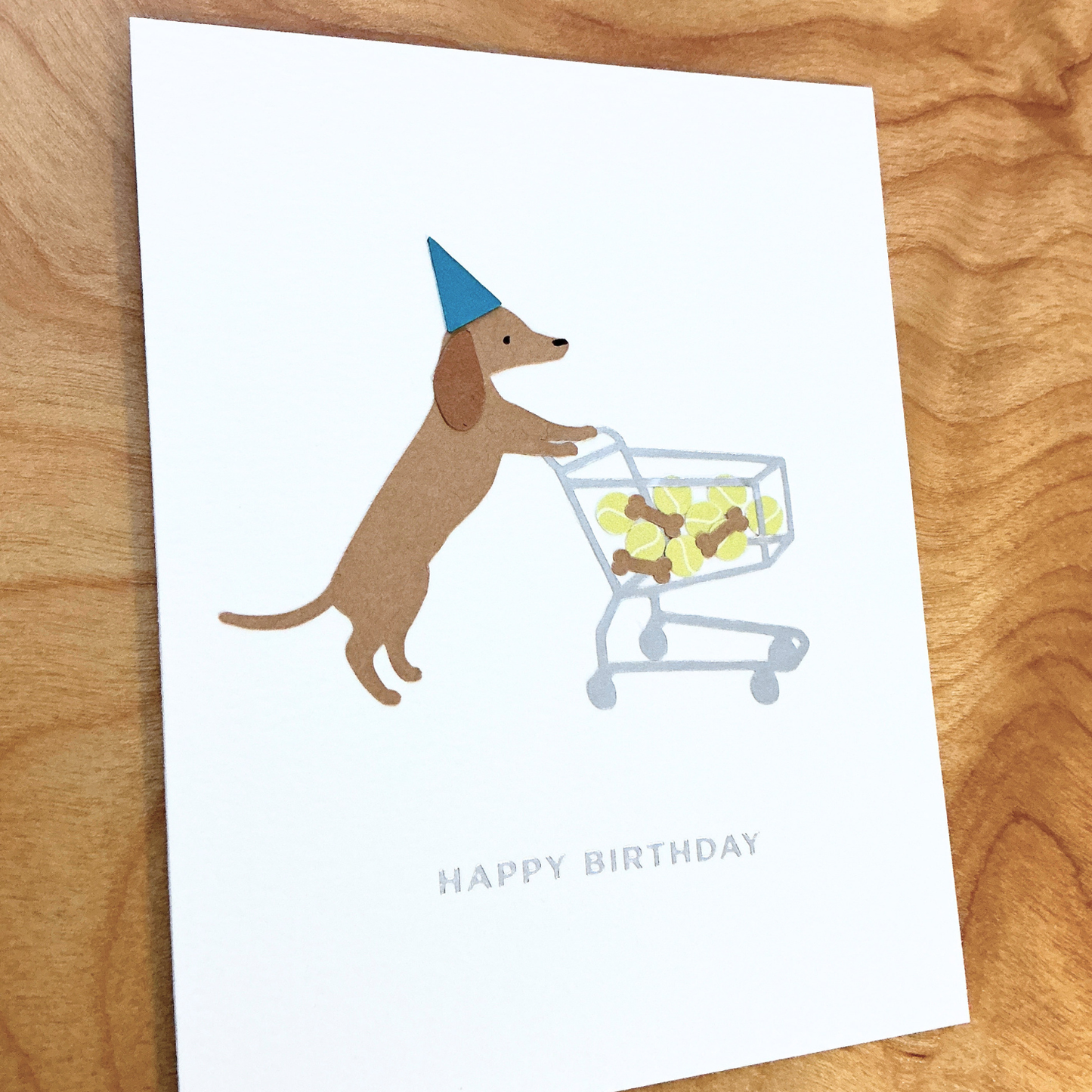 Folded greeting card laying flat on wood surface with a dachshund in a blue party hat pushing a shopping cart full of brown dog treats and tennis balls. Under the dog is silver-colored phrase "Happy Birthday" in foil embossed all caps font. The greeting card design is centered on a white textured card background.