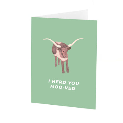 Folded greeting card standing up vertically  with a sage green background featuring a hand-painted style illustration of a longhorn cow with large, curved white horns. below the cow, the phrase "i herd you moo-ved" is written in bold white capital letters. The greeting card is vertically standing up in the center of a white background.