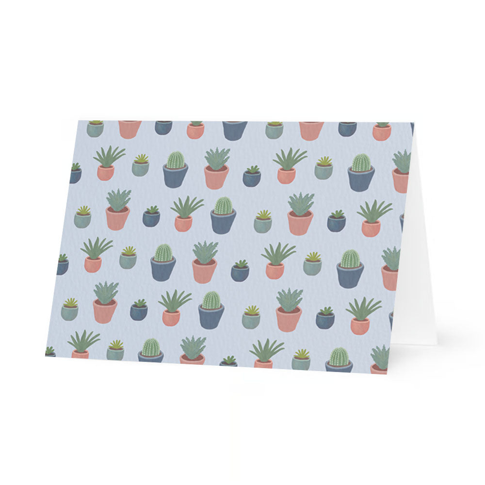 A horizontal, standing folded greeting card shown from a front-facing perspective. The card features a repeating pattern of various illustrated potted plants, including cacti and succulents in shades of green. The plants are housed in small pots of terracotta pink, muted teal, and dark grey, all set against a soft, pale lavender-blue background.