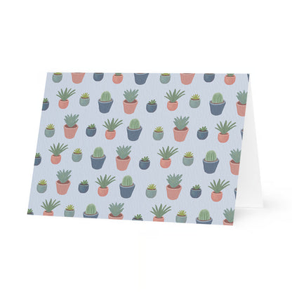A horizontal, standing folded greeting card shown from a front-facing perspective. The card features a repeating pattern of various illustrated potted plants, including cacti and succulents in shades of green. The plants are housed in small pots of terracotta pink, muted teal, and dark grey, all set against a soft, pale lavender-blue background.